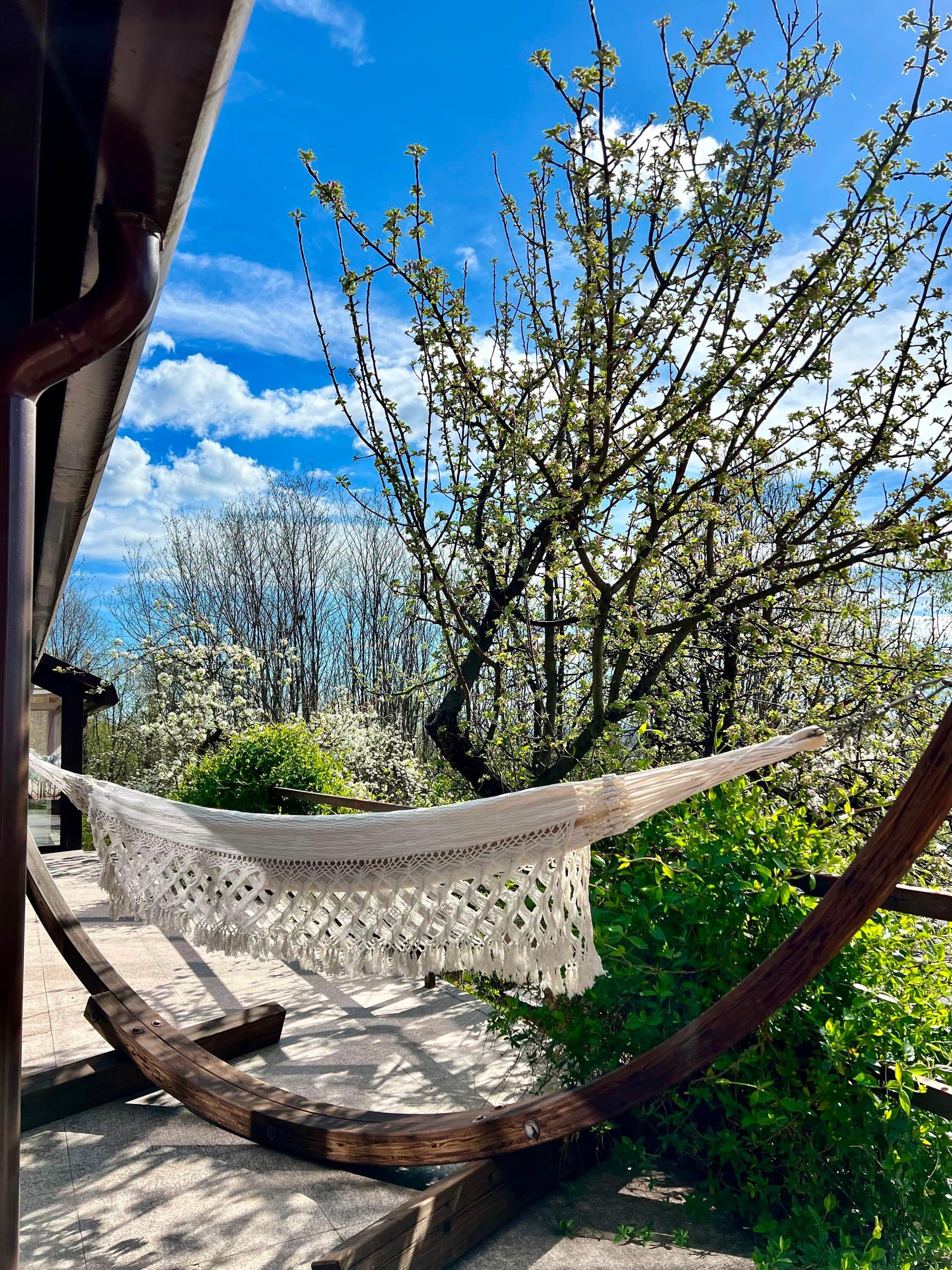 Hamac alb de macrame pe un suport curbat de lemn, pe terasa Hidden Hills, cu pomi înfloriți și cer senin în spate
