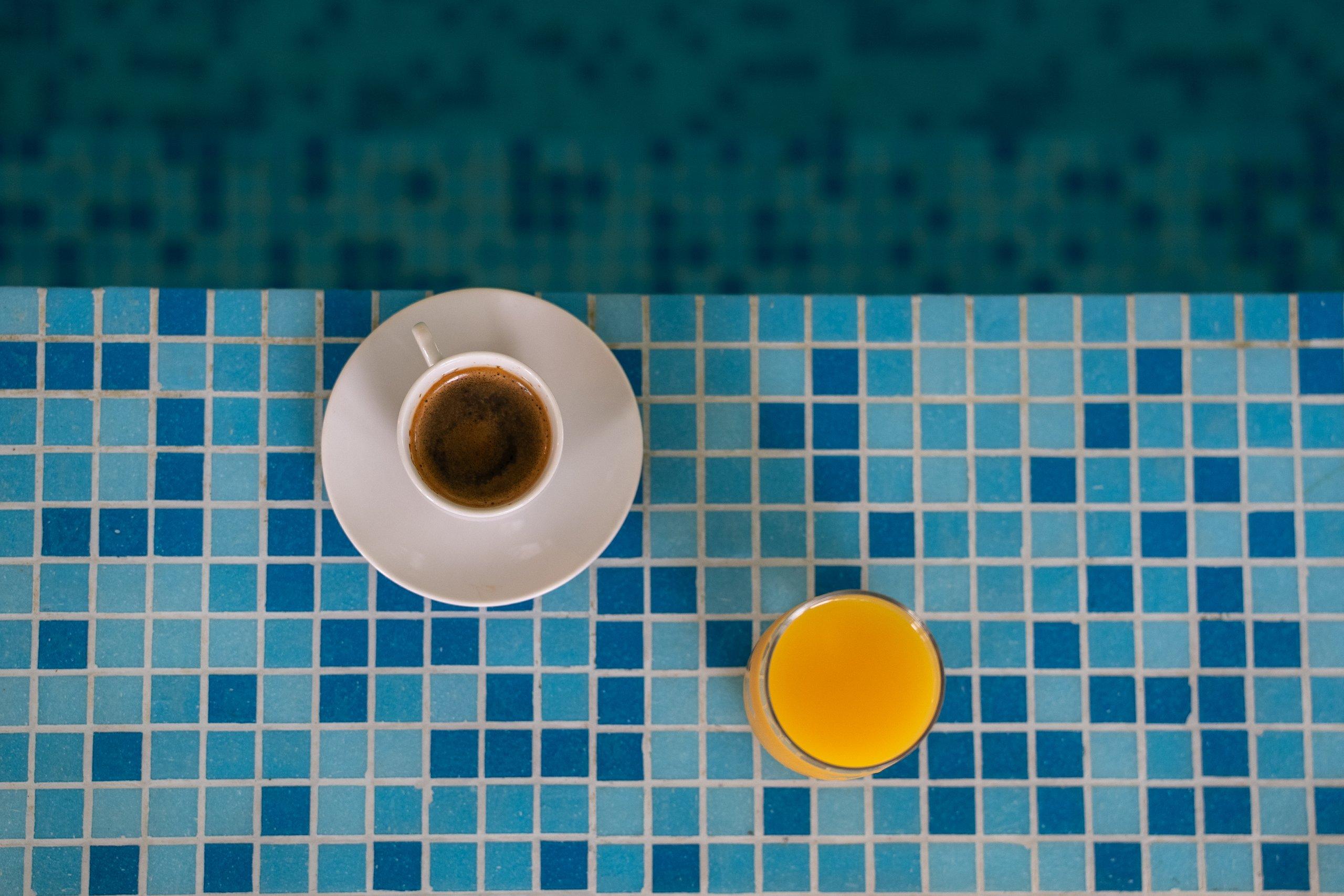 Ceașcă de cafea și un pahar de suc de portocale, pe marginea din mozaic albastru a piscinei interioare